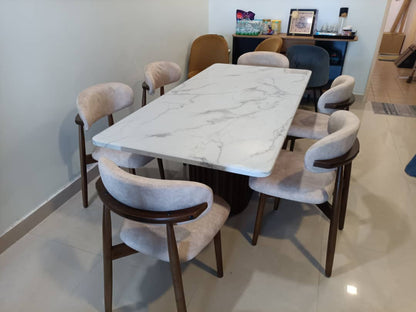 A modern dining set featuring a rectangular white marble-patterned table with a dark wood base, surrounded by six upholstered chairs with beige fabric and dark wood frames, set in a home interior.