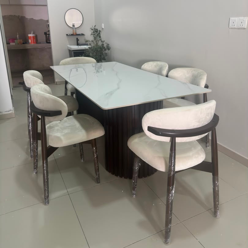 A modern dining set featuring a rectangular white marble-patterned table with a dark fluted pedestal base and six upholstered chairs with cream fabric and dark wood frames, shown in a home interior setting.