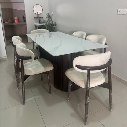 A modern dining set featuring a rectangular white marble-patterned table with a dark fluted pedestal base and six upholstered chairs with cream fabric and dark wood frames, shown in a home interior setting.