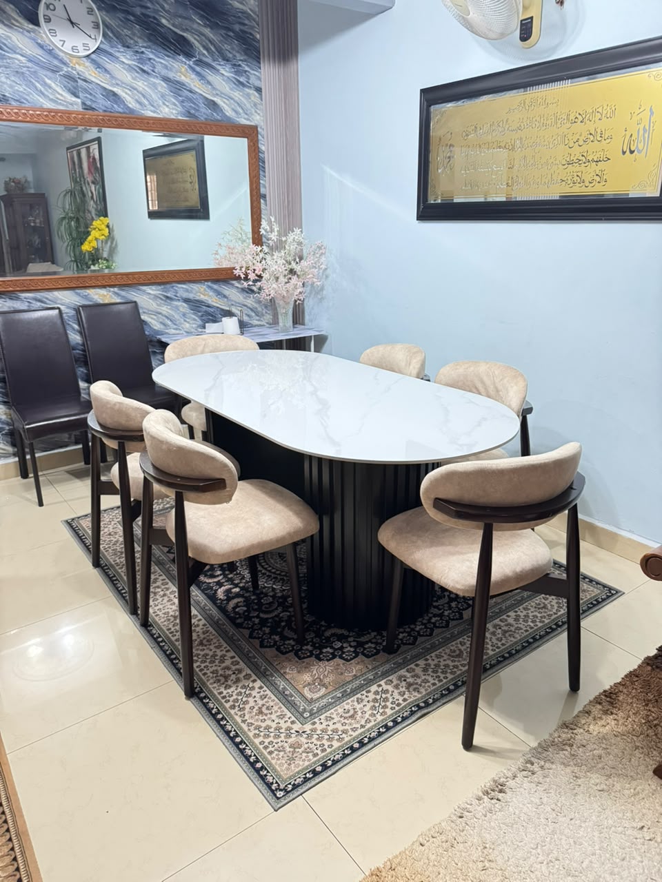 An oval dining table with a white marble-patterned top and a dark fluted pedestal base, surrounded by six upholstered chairs with beige cushions and dark wood frames, set on a patterned rug in a dining room.