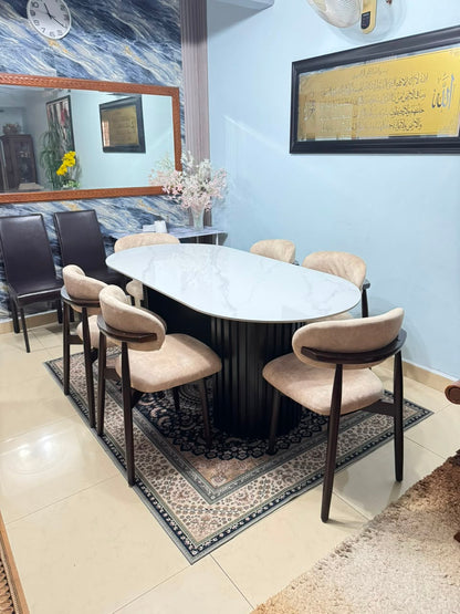An oval dining table with a white marble-patterned top and a dark fluted pedestal base, surrounded by six upholstered chairs with beige cushions and dark wood frames, set on a patterned rug in a dining room.