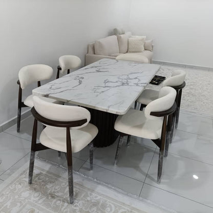 A modern dining set featuring a rectangular white marble-patterned table with a black pedestal base, surrounded by six white upholstered chairs with dark wood armrests and chrome legs, set in a minimalist dining room.