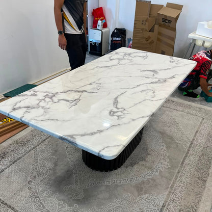 A rectangular coffee table with a white marble-patterned top and a black ribbed pedestal base, photographed in a showroom or warehouse setting on a large patterned rug. A person is partially visible at the upper left, and cardboard boxes and store items are in the background.